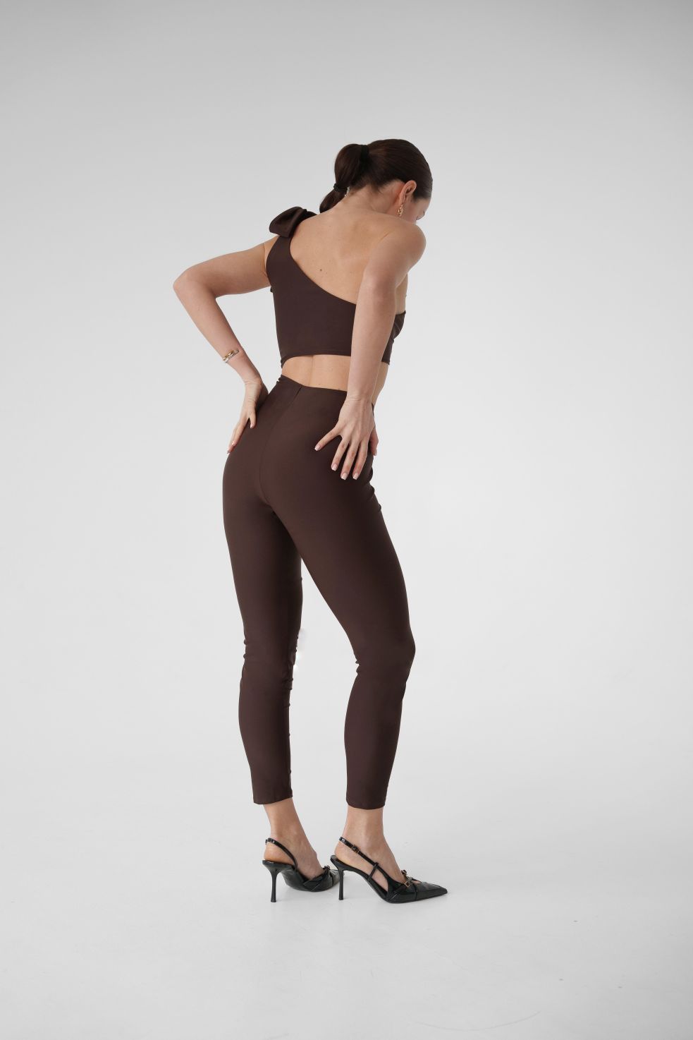 Model wearing Dominica brown one-shoulder jumpsuit with bow strap and leggings