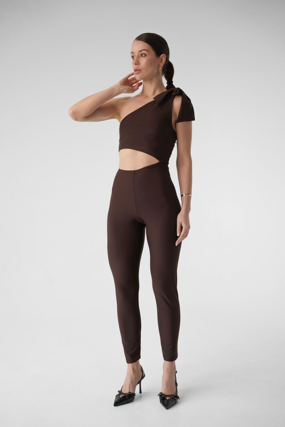 Model wearing Dominica brown one-shoulder jumpsuit with bow strap and leggings